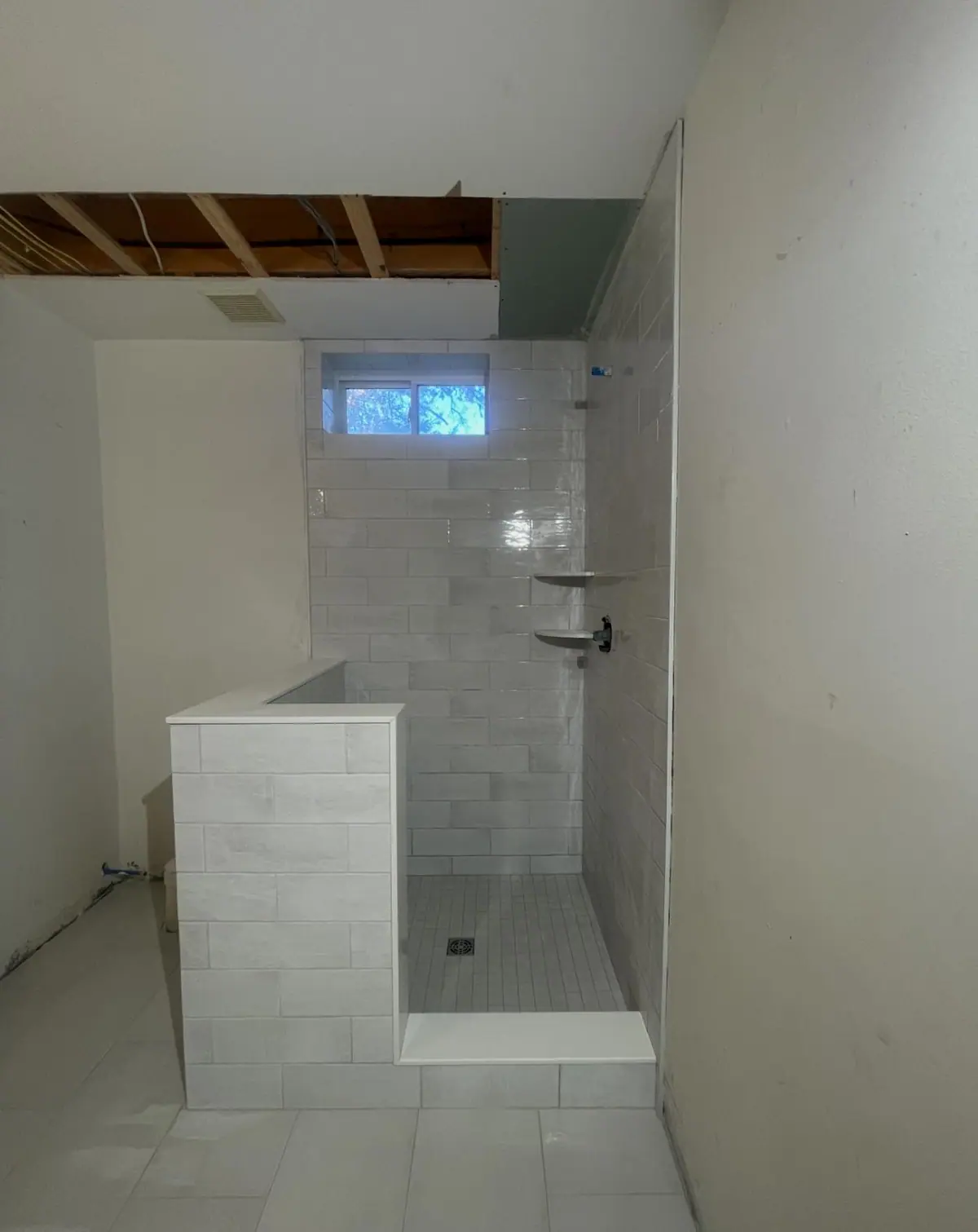 Bathroom with a light gray tiled floor and a walk-in shower behind a tiled half wall with a window