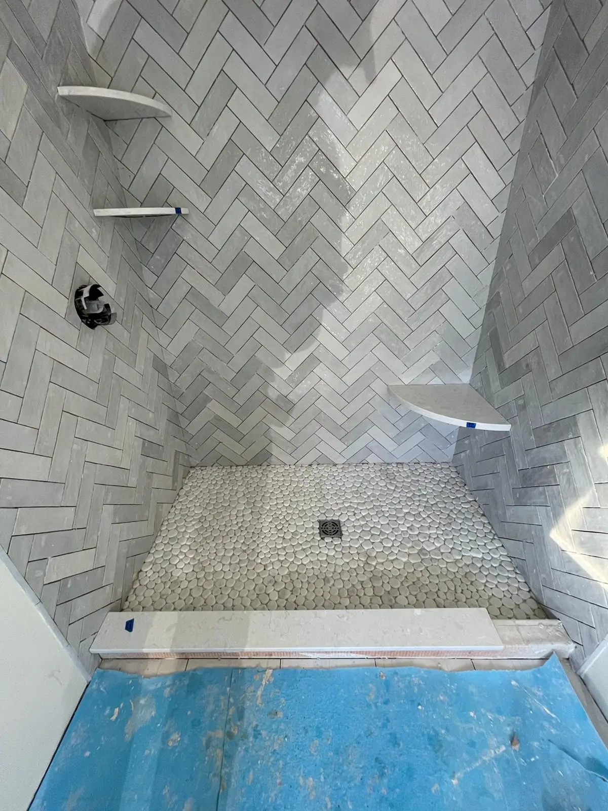 Shower walls in gray herringbone tile with corner shelves and a pebble tile shower floor