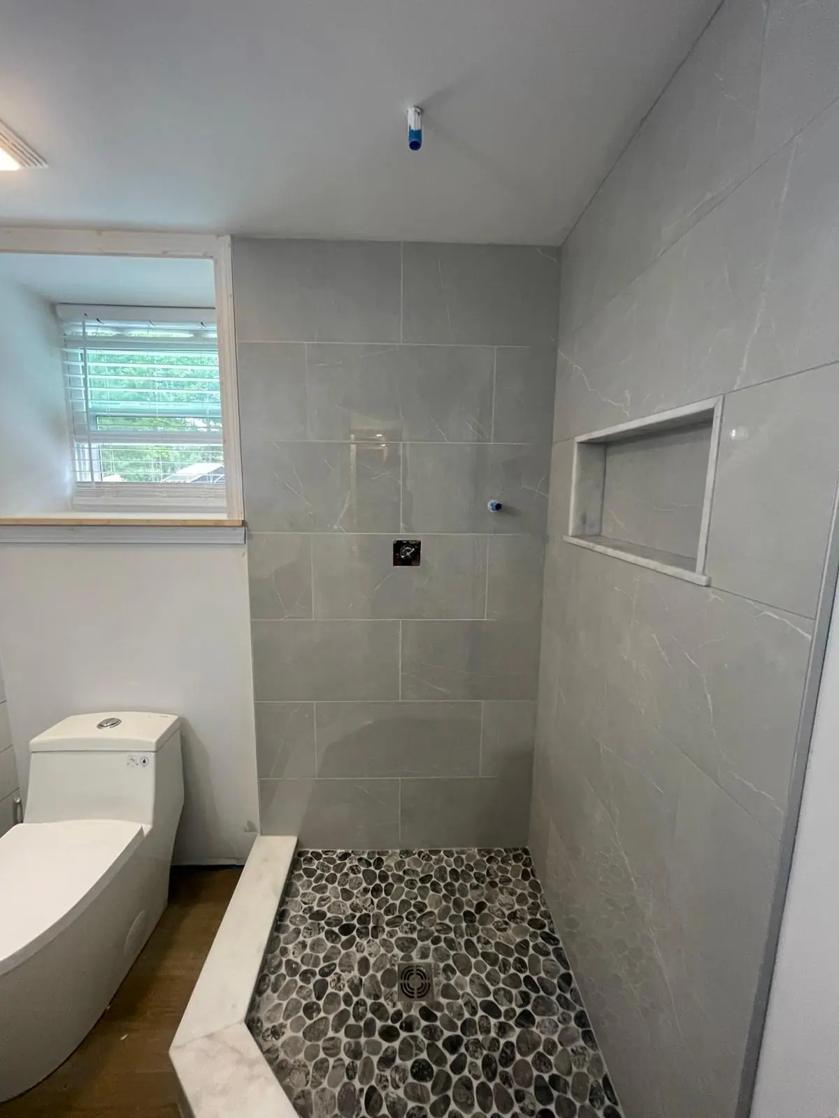 Walk-in shower with light gray wall tile, a recessed niche and a dark pebble tile floor